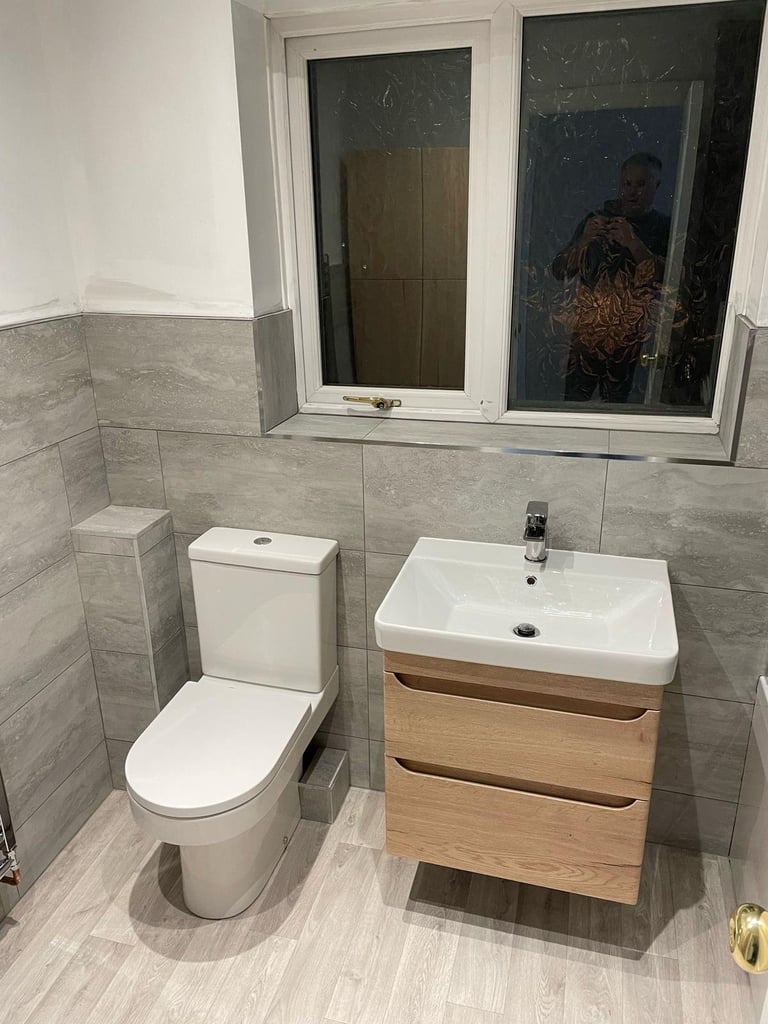 Modern bathroom with gray tile walls, white toilet, wooden vanity with white sink, and window with persons reflection visible