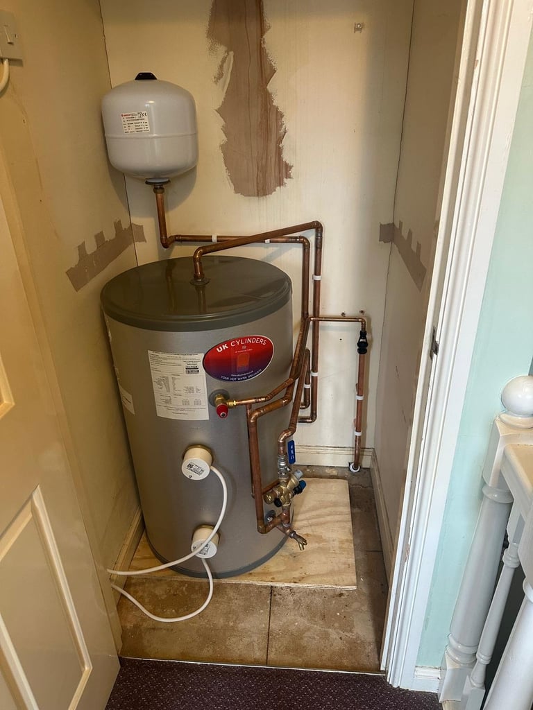 A gray water heater in a utility closet with copper piping, expansion tank above, and white supply line, mounted on tile flooring