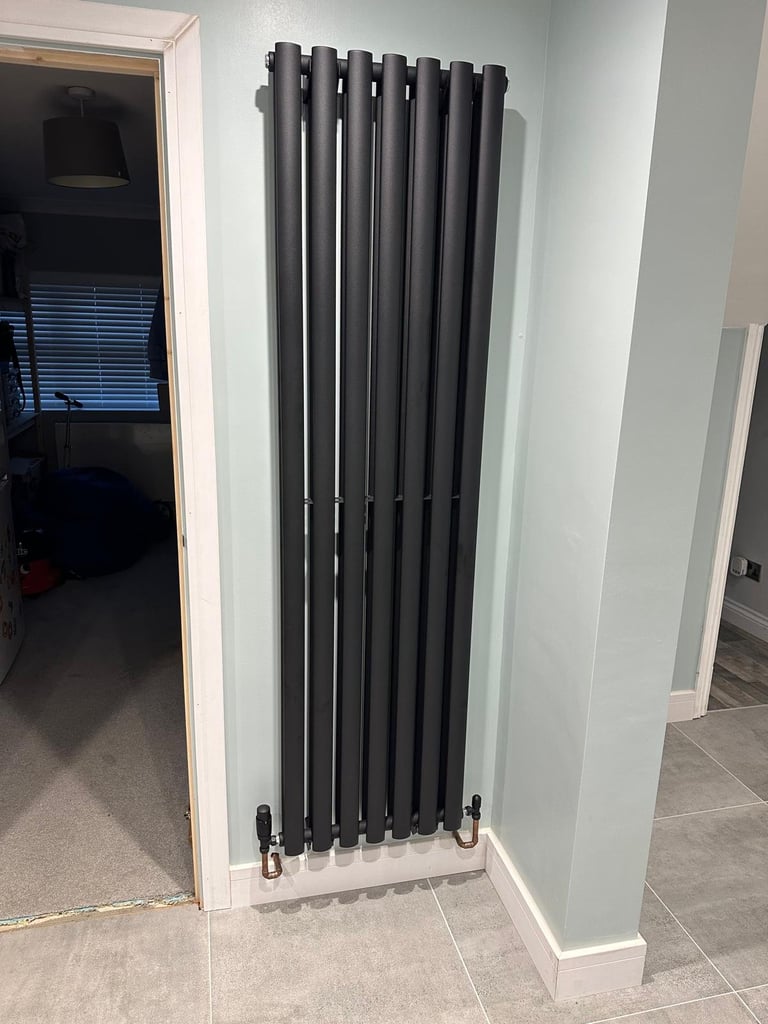 Black vertical radiator mounted on white wall in modern hallway with doorway visible