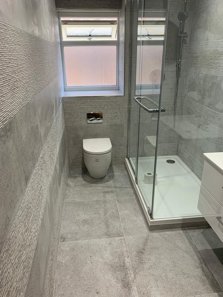 Modern bathroom with white toilet, glass shower enclosure, gray tiled walls and floor, and window with pink sky view