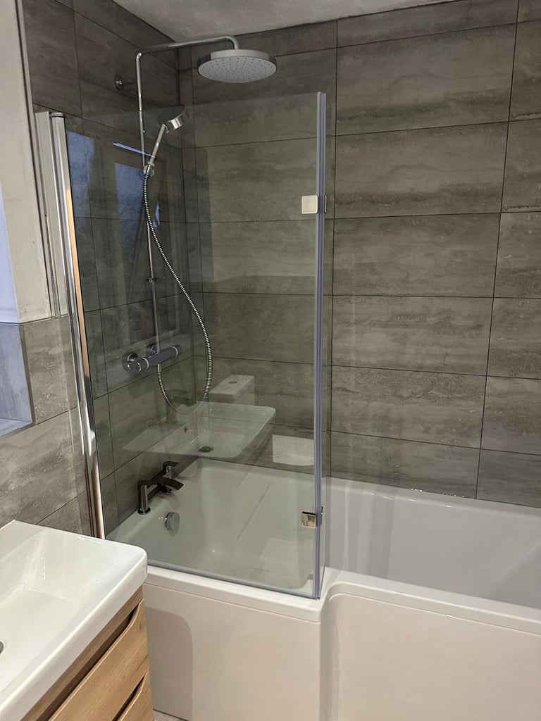 Modern bathroom with frameless glass shower enclosure over white bathtub, gray tile walls, rainfall showerhead, and wooden vanity