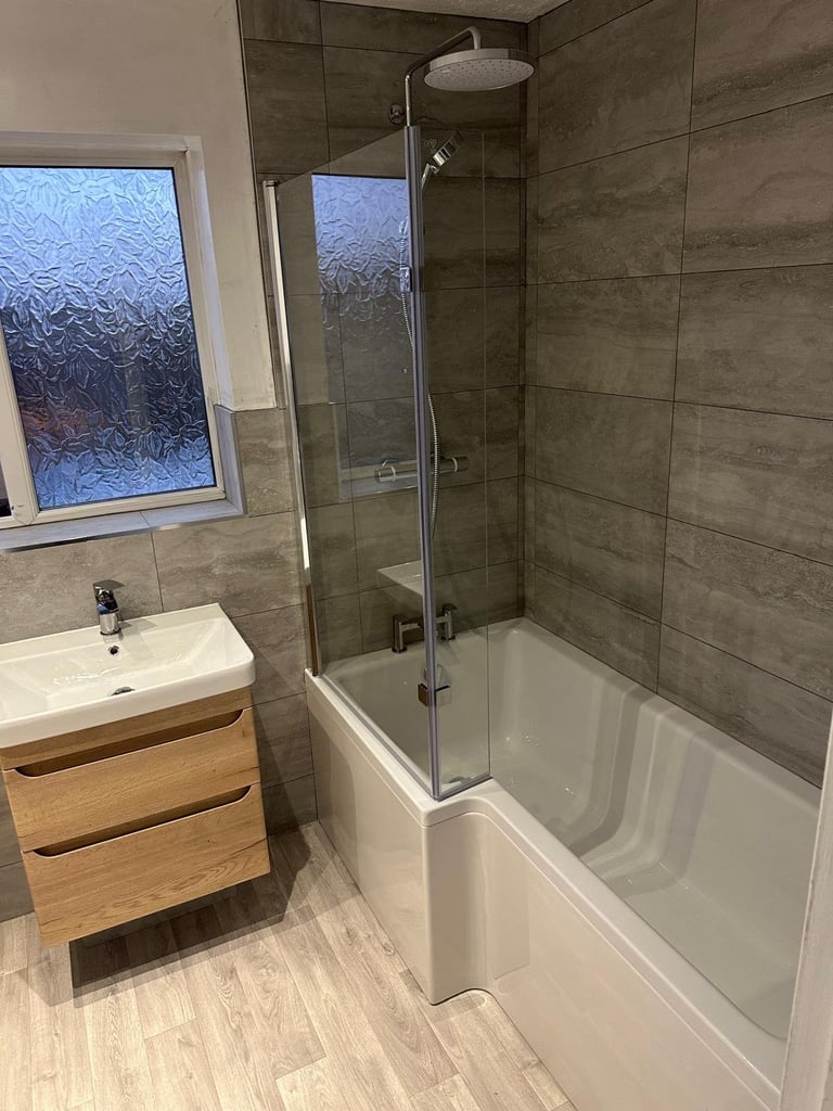 Modern bathroom with gray tile walls, white bathtub with glass shower enclosure, wooden vanity sink, and frosted window