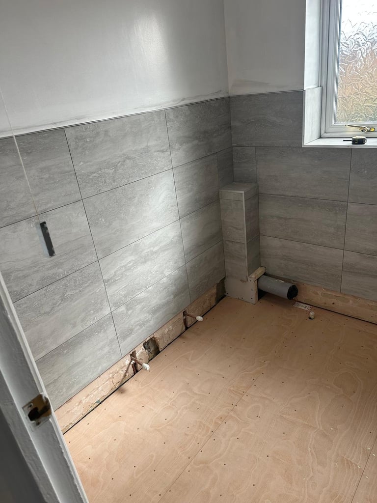 Bathroom under renovation with gray tile walls, white upper walls, plywood floor, and a window