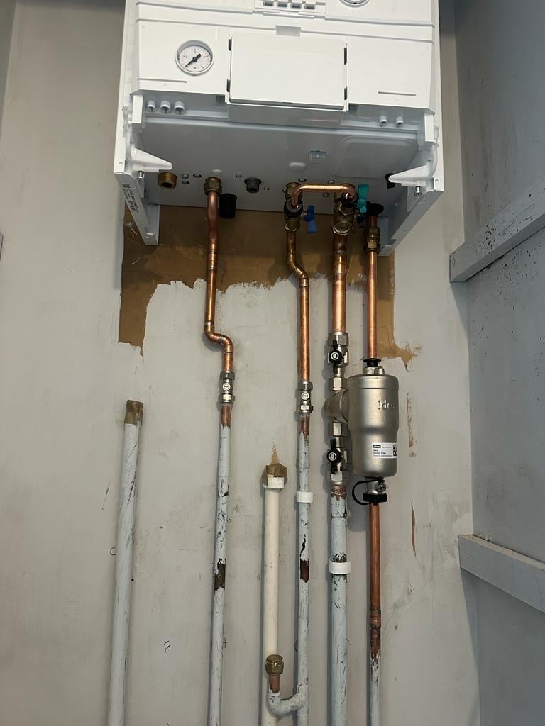 Wall-mounted heating system with exposed copper and white pipes, pressure gauge, and water heater component installed indoors