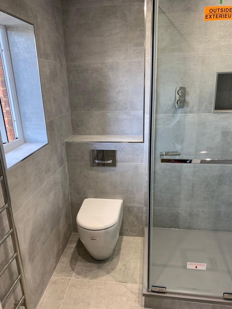 Modern bathroom with white toilet, gray tile walls, glass shower enclosure with metal frame, and small window