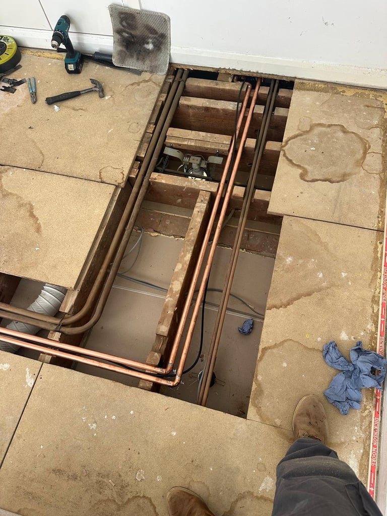 Open floor cavity showing copper pipes and wiring installation with tools and materials surrounding the construction area